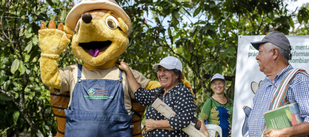 (Left to right) Myriam Contreras Avella, Virginia Montenegro, Jesus Adriano Lancheros, local cacao farmers, at a community event with Armandilla, the armadillo mascot for the Lands for Life program in Meta, Colombia. The armadillo is a representative animal species in Meta department. The community selected this female mascot to highlight the importance of integrating biodiversity (fauna and flora) into their production systems, since it is an endangered species. Its original name is Amandilla but the community calls it Armandilla because they associate it with the species.