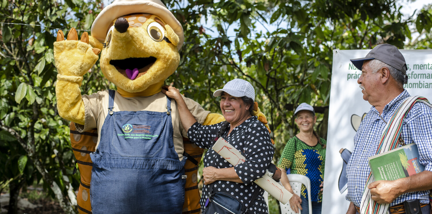 (Left to right) Myriam Contreras Avella, Virginia Montenegro, Jesus Adriano Lancheros, local cacao farmers, at a community event with Armandilla, the armadillo mascot for the Lands for Life program in Meta, Colombia. The armadillo is a representative animal species in Meta department. The community selected this female mascot to highlight the importance of integrating biodiversity (fauna and flora) into their production systems, since it is an endangered species. Its original name is Amandilla but the community calls it Armandilla because they associate it with the species.